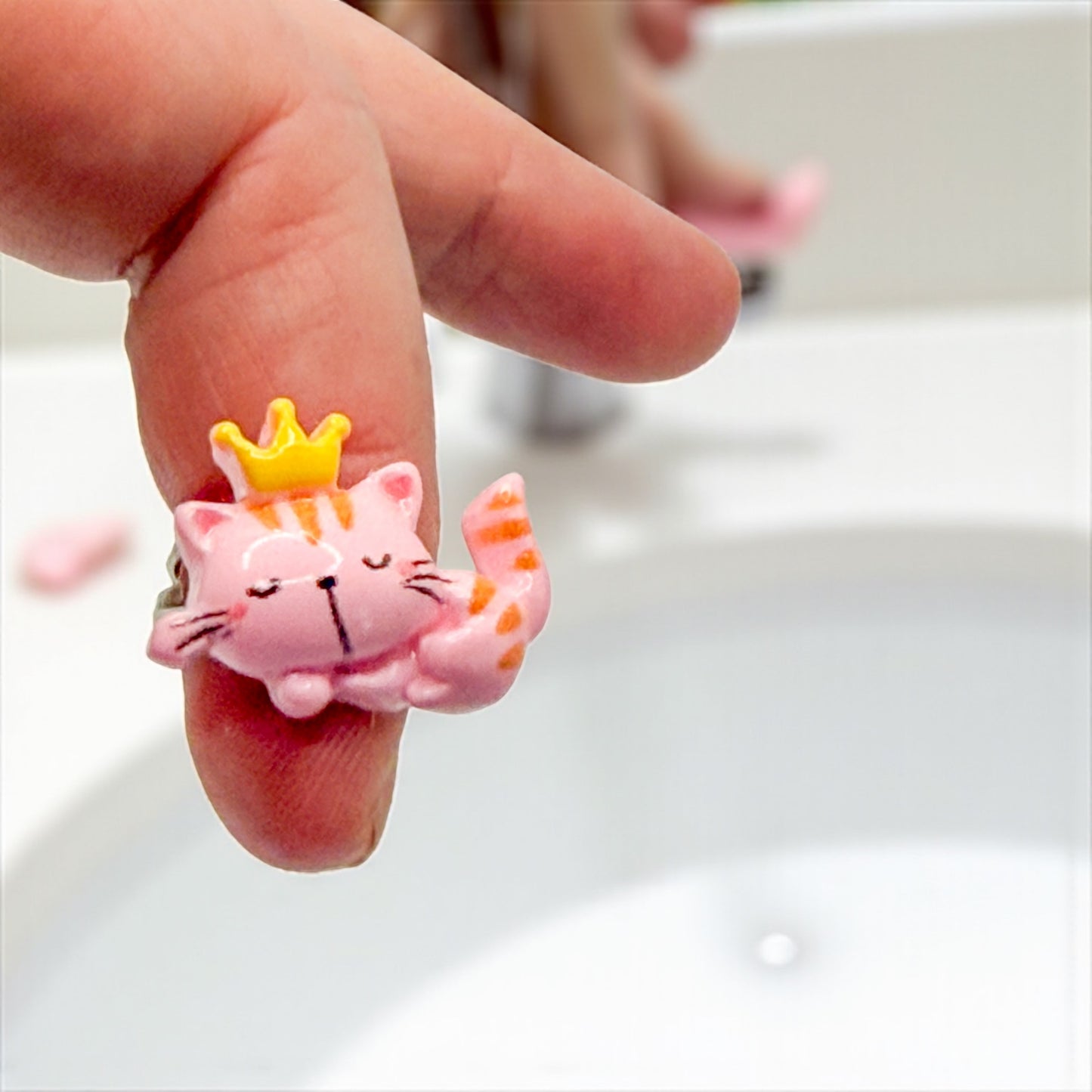 Pink cat figurine with a yellow crown on a person's finger against a white background