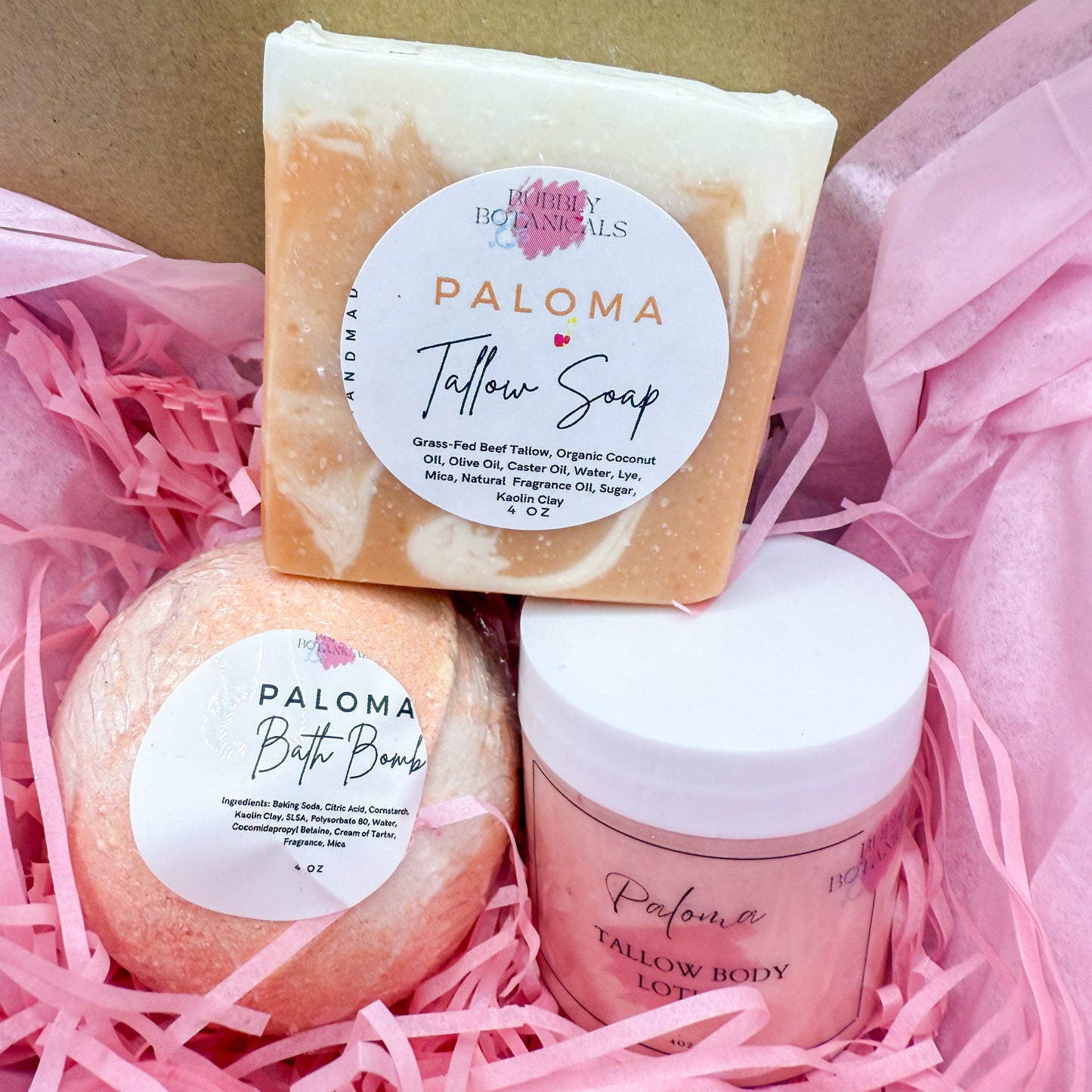 Three Paloma products including a soap bar, bath bomb, and body scrub in pink shredded paper.
