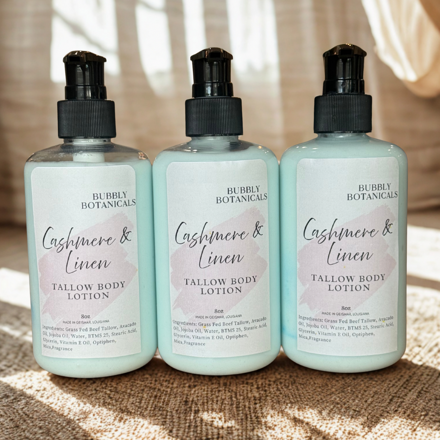 Three bottles of Bubbly Botanicals Cashmere & Linen Tallow Body Lotion on a textured surface with sunlight filtering through curtains.