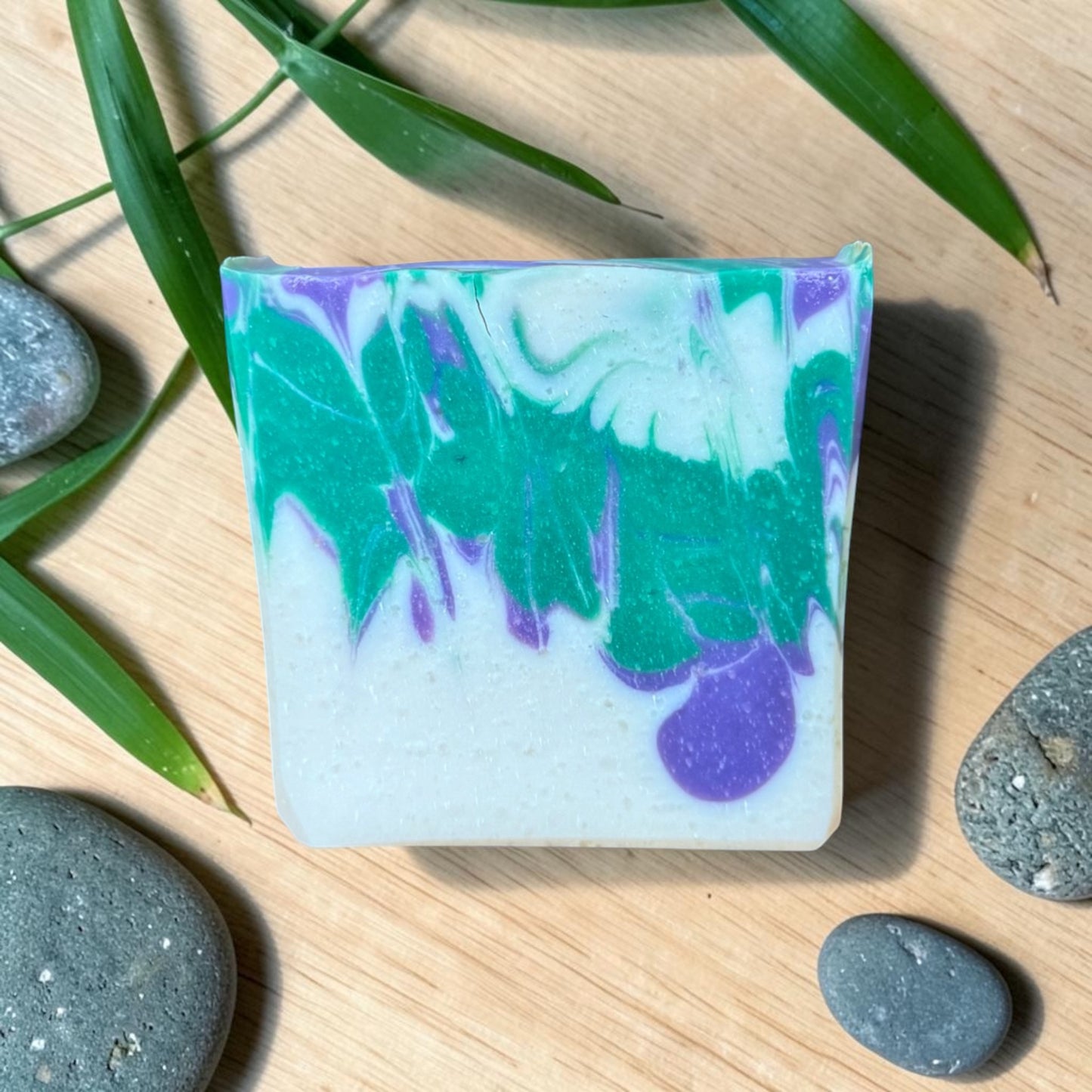Colorful soap bar with green, purple, and white swirls on a wooden surface with stones and leaves.