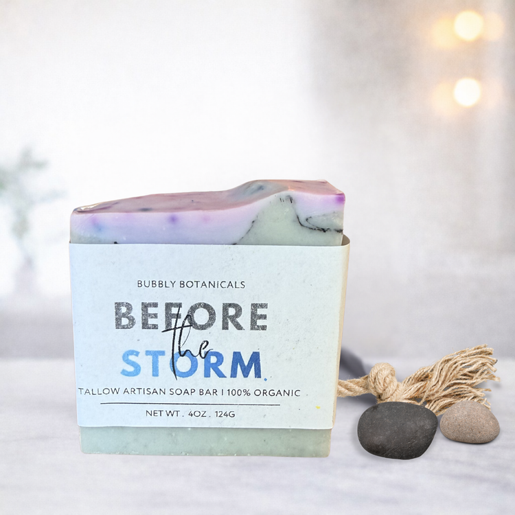 A handmade tallow soap bar with a white paper label reading 'Bubbly Botanicals Before the Storm,' styled on a light surface with small stones and a tassel ornament against a softly blurred warm background.