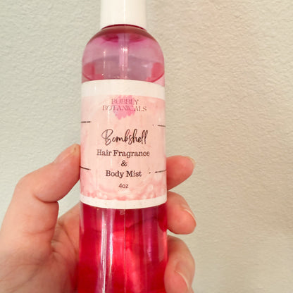 Hand holding a bottle of 'Bombshell' hair fragrance and body mist against a plain background