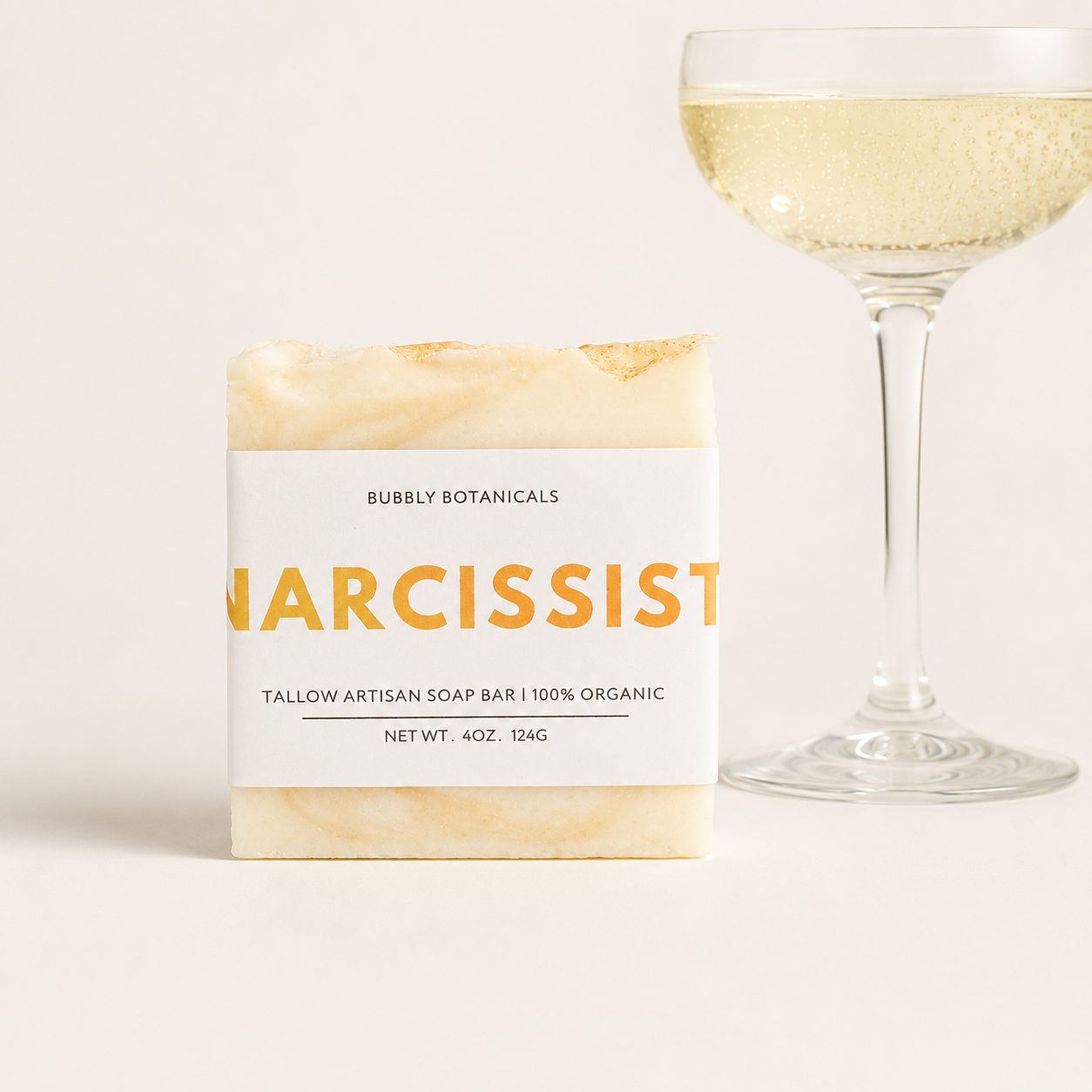 A bar of artisanal tallow soap labeled 'Narcissist (type)' by Bubbly Botanicals, shown next to a champagne glass filled with bubbly liquid on a light beige background.