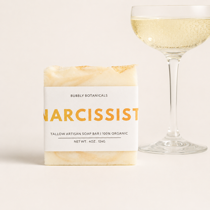 A bar of artisanal tallow soap labeled 'Narcissist (type)' by Bubbly Botanicals, shown next to a champagne glass filled with bubbly liquid on a light beige background.