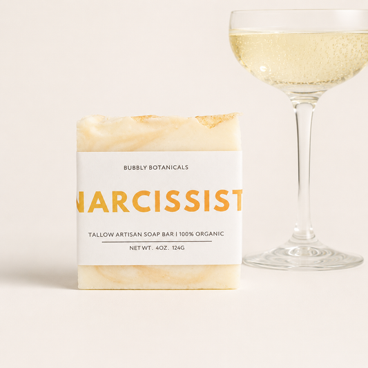 A bar of artisanal tallow soap labeled 'Narcissist (type)' by Bubbly Botanicals, shown next to a champagne glass filled with bubbly liquid on a light beige background.