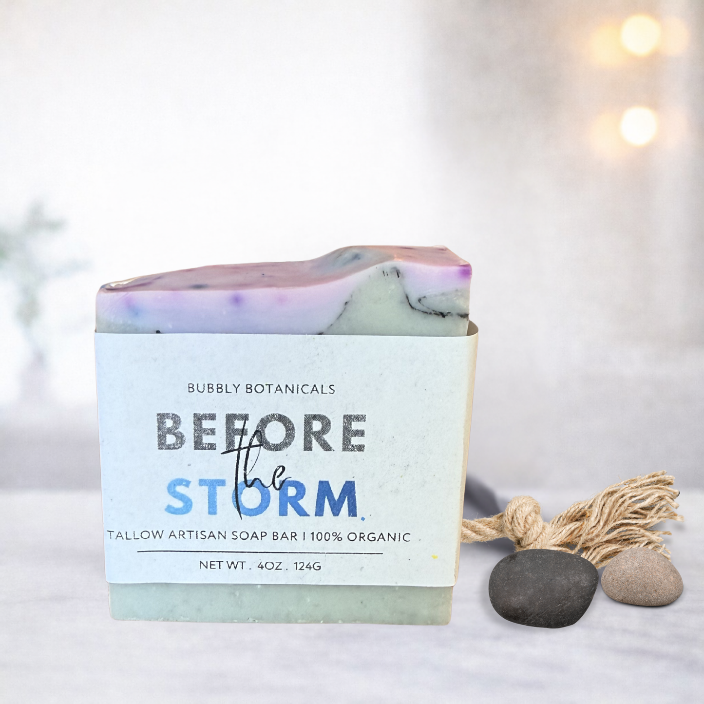 A handmade tallow soap bar with a white paper label reading 'Bubbly Botanicals Before the Storm,' styled on a light surface with small stones and a tassel ornament against a softly blurred warm background.