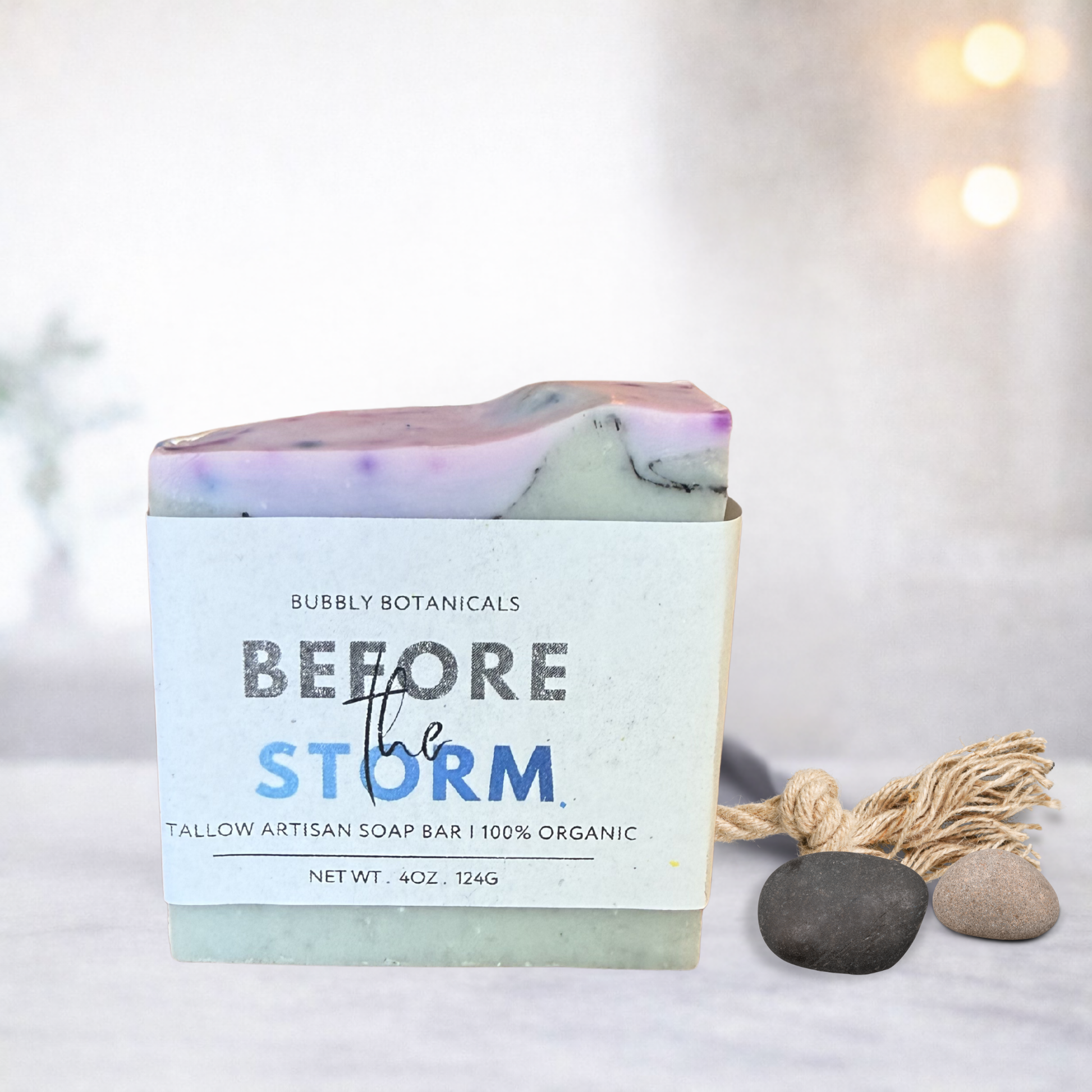 A handmade tallow soap bar with a white paper label reading 'Bubbly Botanicals Before the Storm,' styled on a light surface with small stones and a tassel ornament against a softly blurred warm background.