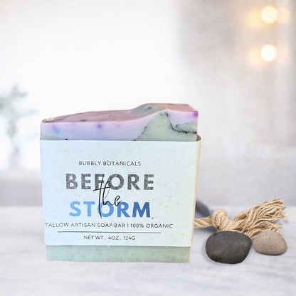 A handmade tallow soap bar with a white paper label reading 'Bubbly Botanicals Before the Storm,' styled on a light surface with small stones and a tassel ornament against a softly blurred warm background.