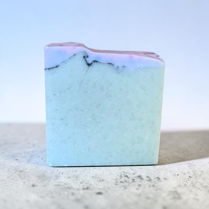 A rectangular bar of handmade tallow soap with a light blue body, subtle white speckles, and a top layer featuring a wavy pattern of white and lavender-purple with thin dark veins, standing on a textured gray surface against a soft blue gradient background.