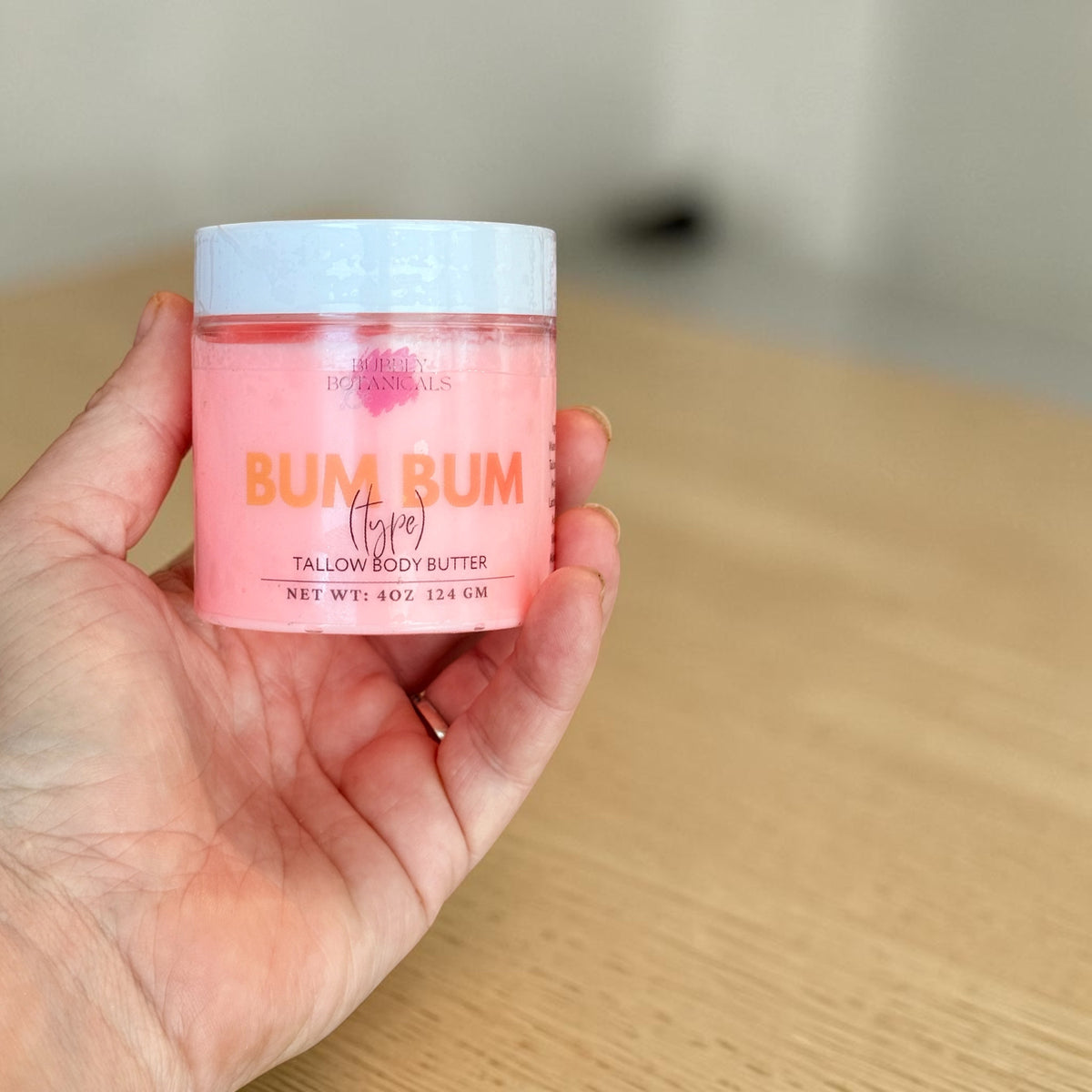 A hand holding a small jar of Bum Bum Tallow Body Butter. The jar is filled with a pink body butter and has a white lid with orange and black text.