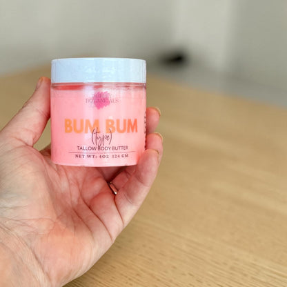 A hand holding a small jar of Bum Bum Tallow Body Butter. The jar is filled with a pink body butter and has a white lid with orange and black text.