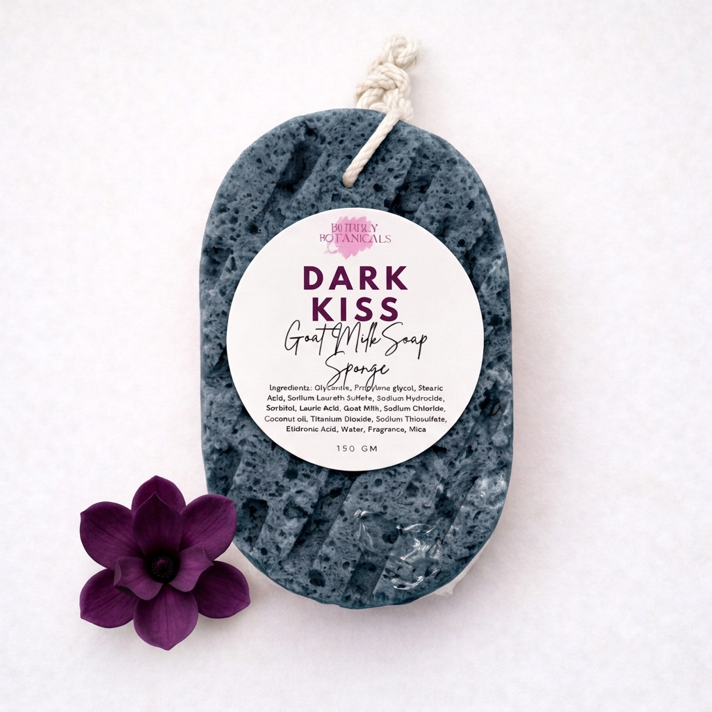 A dark slate gray goat milk soap sponge with a white hanging tag reading 'DARK KISS GOAT milk Soap Sponge' and a purple flower prop on a white background.