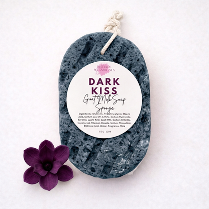 A dark slate gray goat milk soap sponge with a white hanging tag reading 'DARK KISS GOAT milk Soap Sponge' and a purple flower prop on a white background.