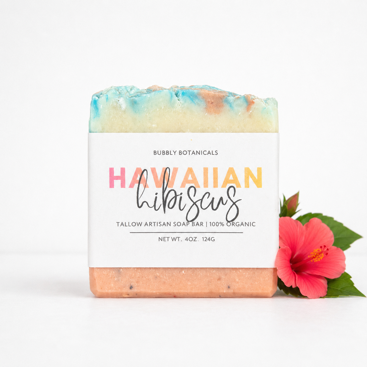 Bar of hawaiian hibiscus grass-fed tallow soap with a colorful top and 'Hawaiian Hibiscus' label, accompanied by a pink flower on a white background.