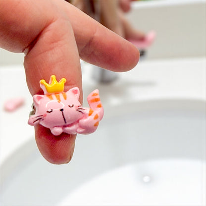 Pink cat figurine with a yellow crown on a person's finger against a white background