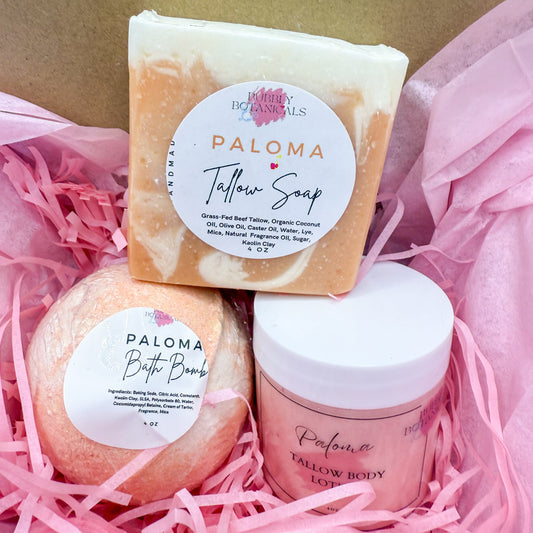Three Paloma products including a soap bar, bath bomb, and body scrub in pink shredded paper.