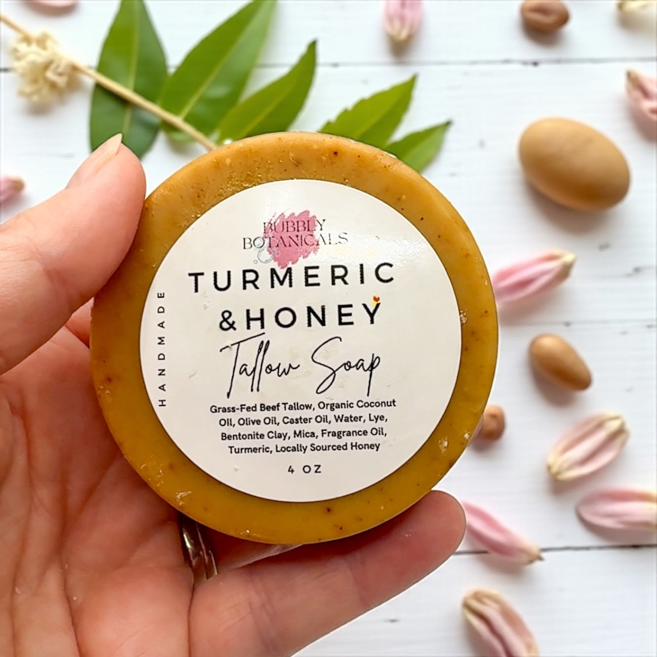 Hand holding a tallow soap bar labeled 'Turmeric & Honey' by Bubly Botanicals on a white background with leaves and flowers.