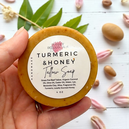 Hand holding a tallow soap bar labeled 'Turmeric & Honey' by Bubly Botanicals on a white background with leaves and flowers.