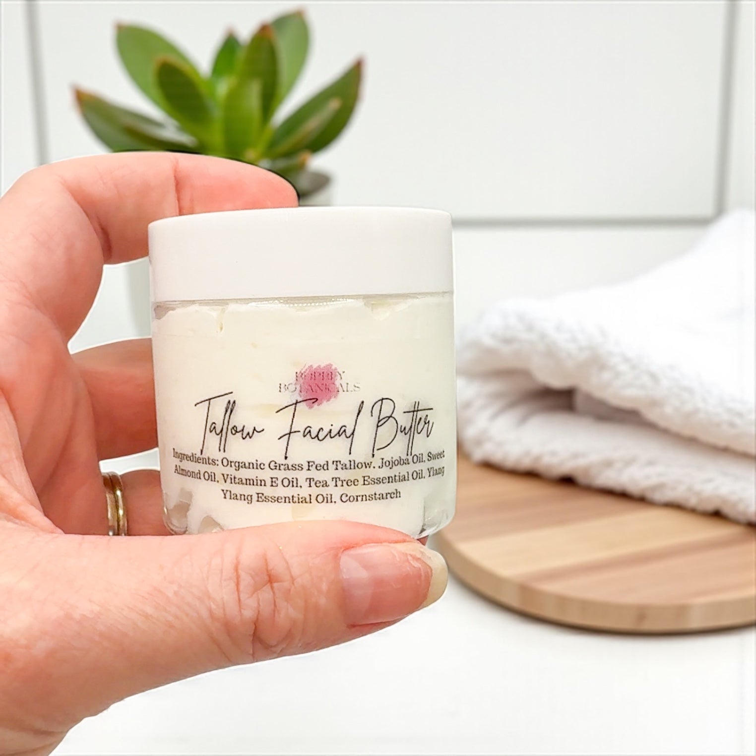 Hand holding a jar of Tallow Facial Butter with a plant and towel in the background
