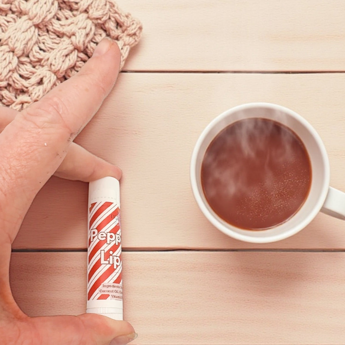Hand holding a lip balm next to a cup of hot chocolate on a wooden surface