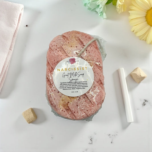 Pink bath sponge with 'Narcissist' label on a white surface with flowers and a towel.