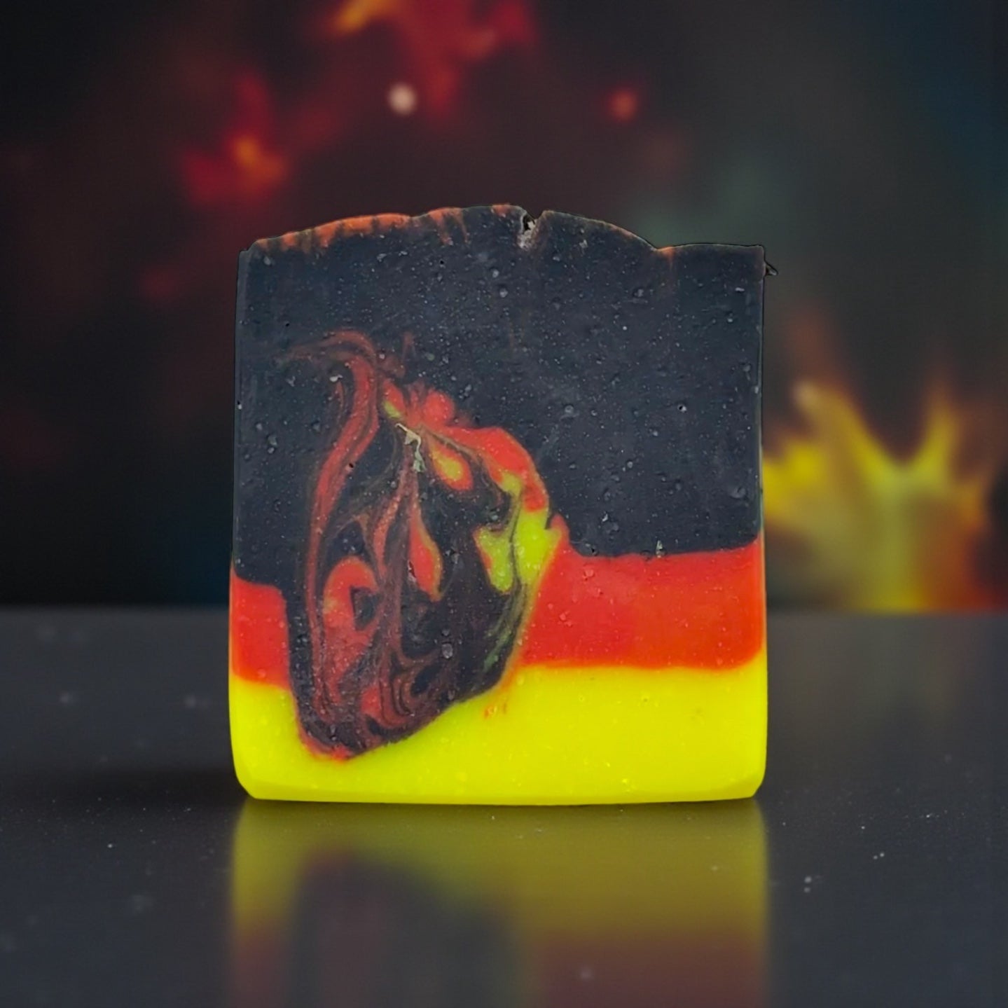 Colorful soap bar with black, red, and yellow layers on a reflective surface with a fiery background.