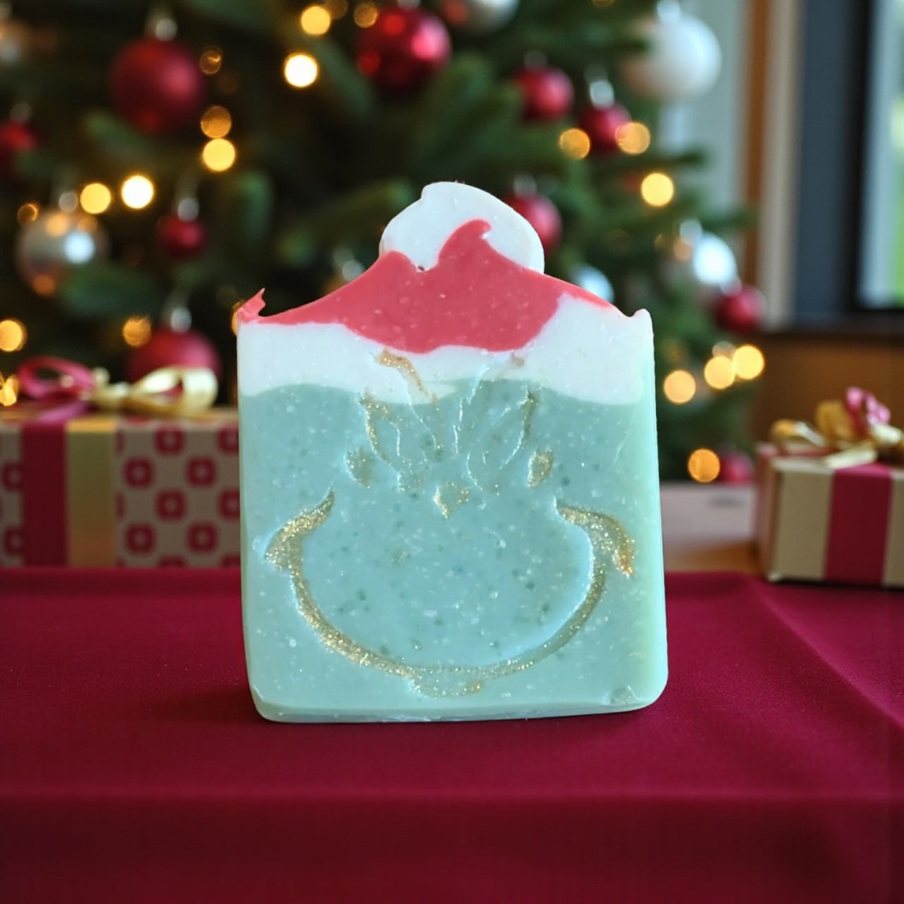 Decorative soap with a Christmas tree design on a red surface, with a blurred Christmas tree and presents in the background.
