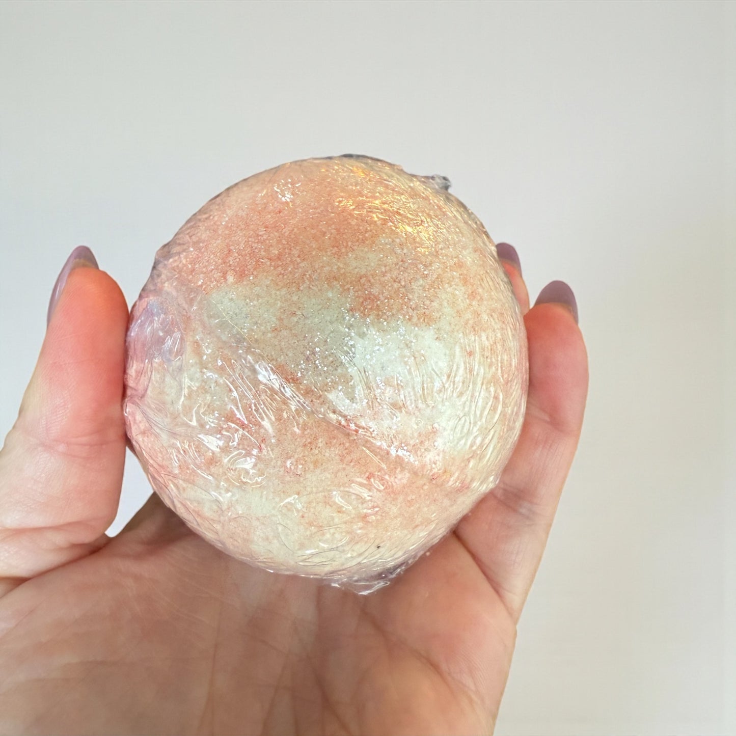 Hand holding a round, pinkish-brown bath bomb against a plain background