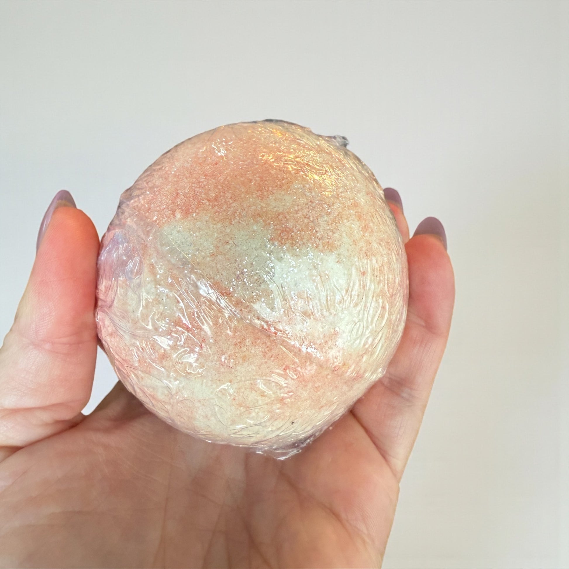 Hand holding a round, pinkish-brown bath bomb against a plain background