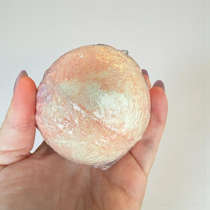 Hand holding a round, pinkish-brown bath bomb against a plain background