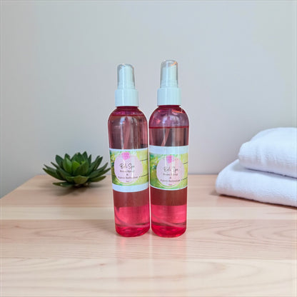 Two spray bottles with pink liquid on a wooden surface with a plant and towels in the background.
