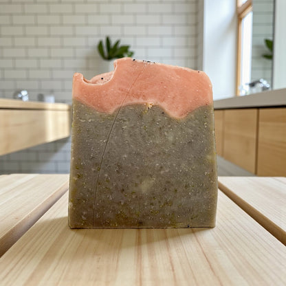 Bar of soap with a pink top and brownish bottom on a wooden surface, kitchen in the background