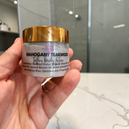 A hand holding a jar of Mahogany Teakwood Tallow Body lotion, showing the white jar with a metallic gold lid against a blurred bathroom background.