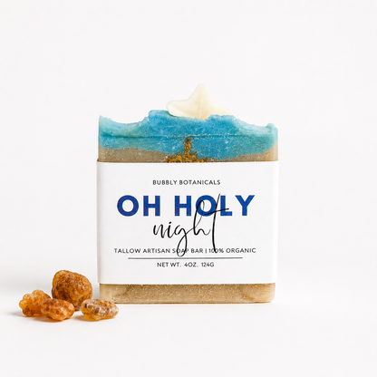 A rectangular artisan tallow soap bar wrapped in a white paper label reading 'OH HOLY night' by Bubbly Botanicals, with a blue and beige marbled top and small amber pieces placed in front on a white background.