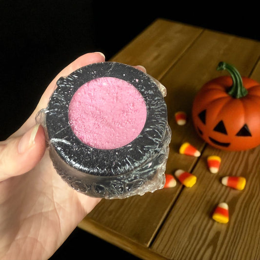 Pink cauldron bath bomb wrapped in black plastic, held by a hand on a wooden surface with Halloween decorations.