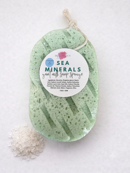 A sea minerals goat milk soap sponge in a light green porous sponge shape, hanging from a twine loop with a white circular label. A small pile of white bath salts sits to the lower left on a white textured background.
