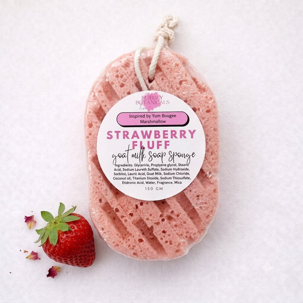 A strawberry-fluff scented goat milk soap sponge wrapped in clear plastic with a white hanging string and a white label. A fresh strawberry and a few petals are placed beside it on a white textured background.