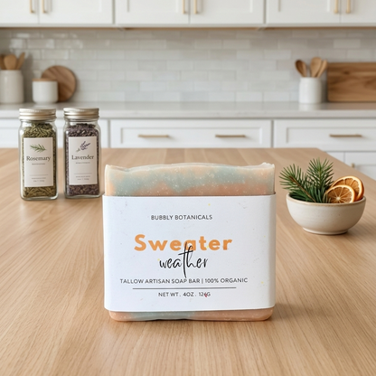 A packaged artisan soap bar labeled 'Sweeter weather' by Bubbly Botanicals sits on a wooden countertop in a bright kitchen setting. Two glass spice jars and a small bowl with dried citrus and greenery are visible in the background.