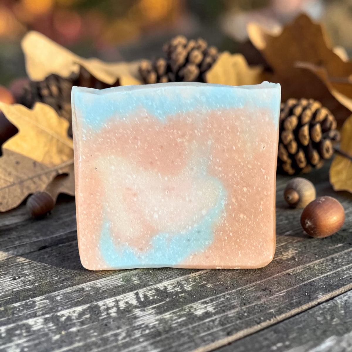 A rectangular bar of handmade tallow soap in the sweater weather scent with a marbled swirl of light blue and peach-pink, placed on a rustic dark wooden surface surrounded by autumn leaves, pinecones, and acorns.