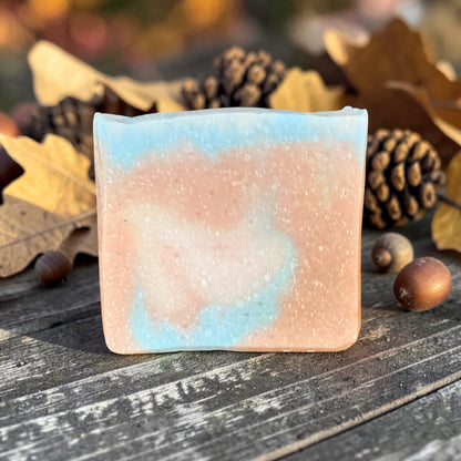 A rectangular bar of handmade tallow soap in the sweater weather scent with a marbled swirl of light blue and peach-pink, placed on a rustic dark wooden surface surrounded by autumn leaves, pinecones, and acorns.
