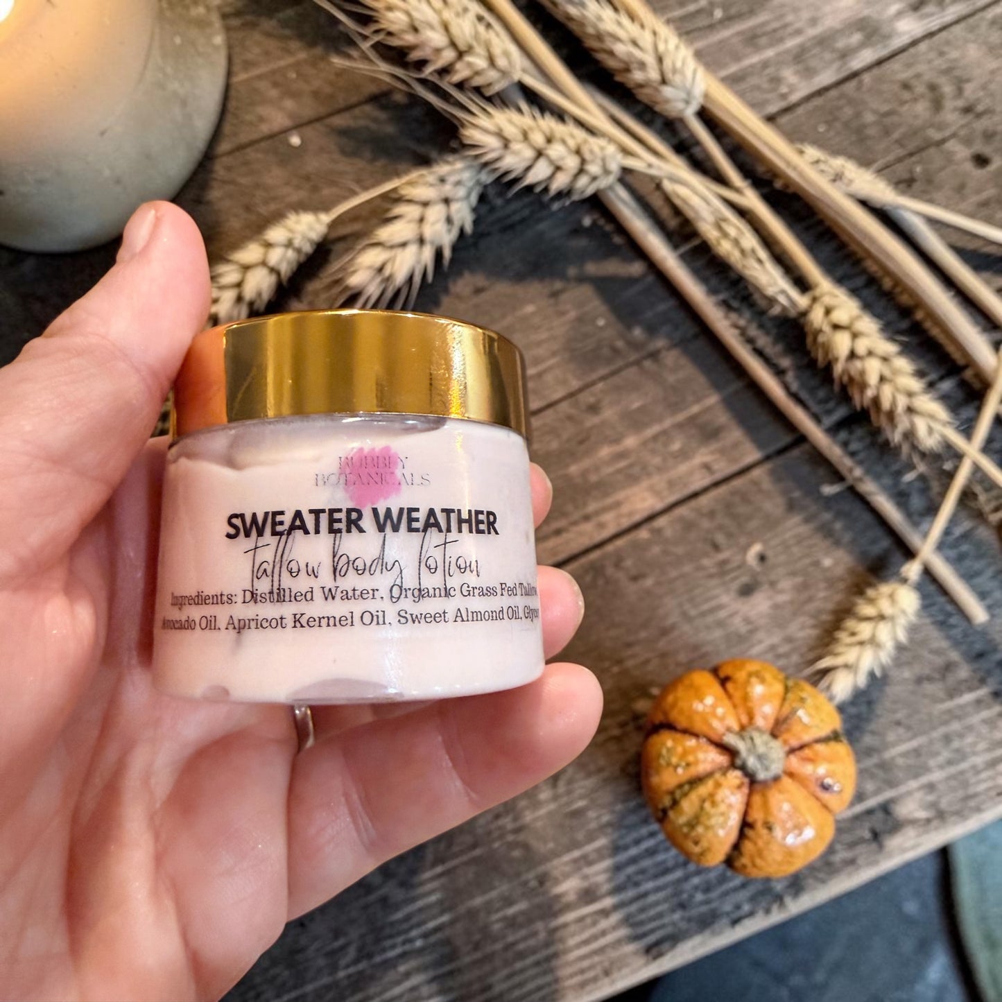 A hand holds a jar of 'Sweater Weather' tallow body lotion. The jar has a gold lid and a pale pink/cream-colored body, set against a rustic wooden background decorated with dried wheat stalks and a small orange pumpkin.