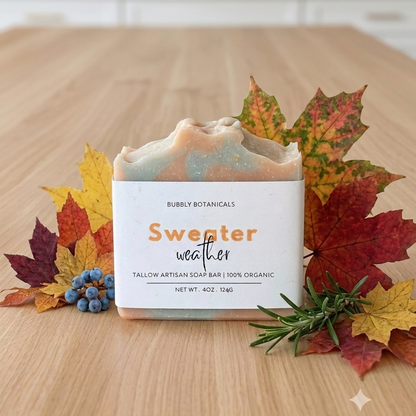 A rectangular bar of handmade tallow soap labeled 'Sweater weather' by Bubble Botanicals, styled on a wooden surface with autumn leaves, blue berries, and rosemary sprigs.