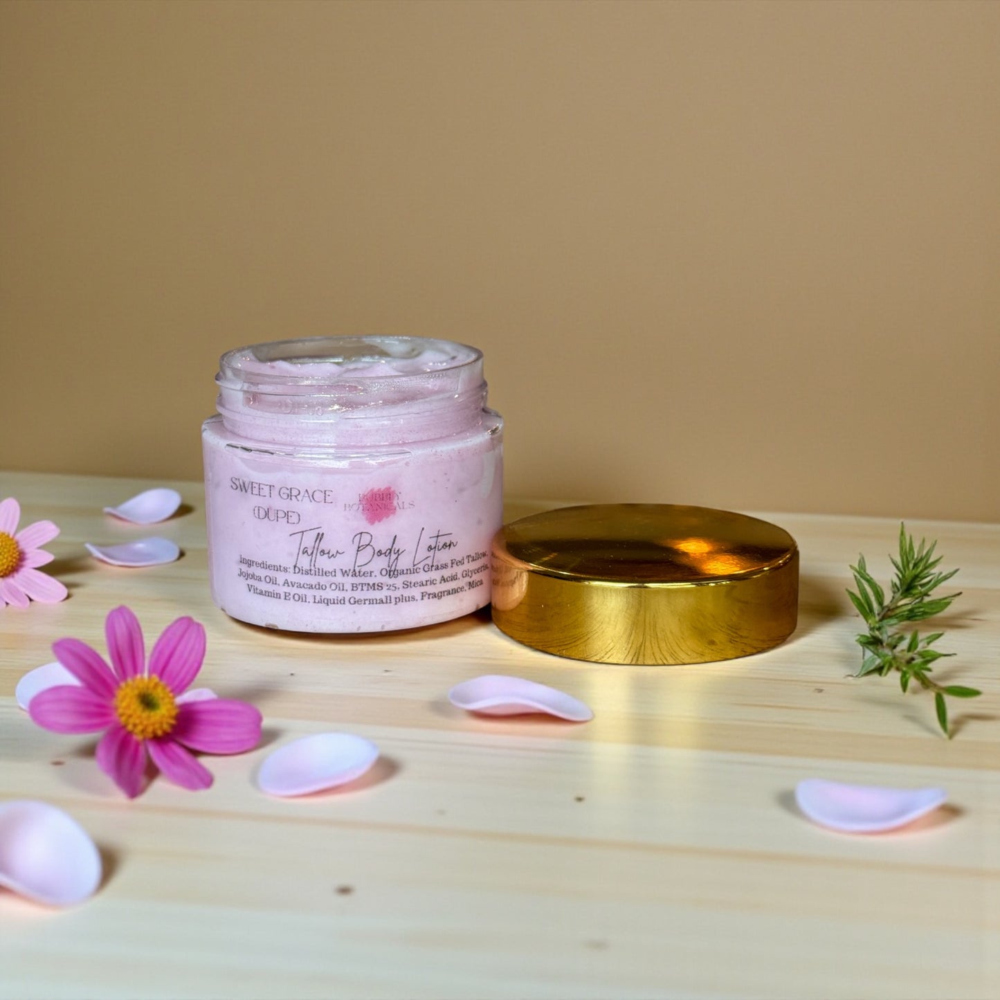 A jar of Sweet Grace Type Tallow Body Lotion with its gold lid beside it, styled on a light wood surface with pink flowers and petals against a beige background.