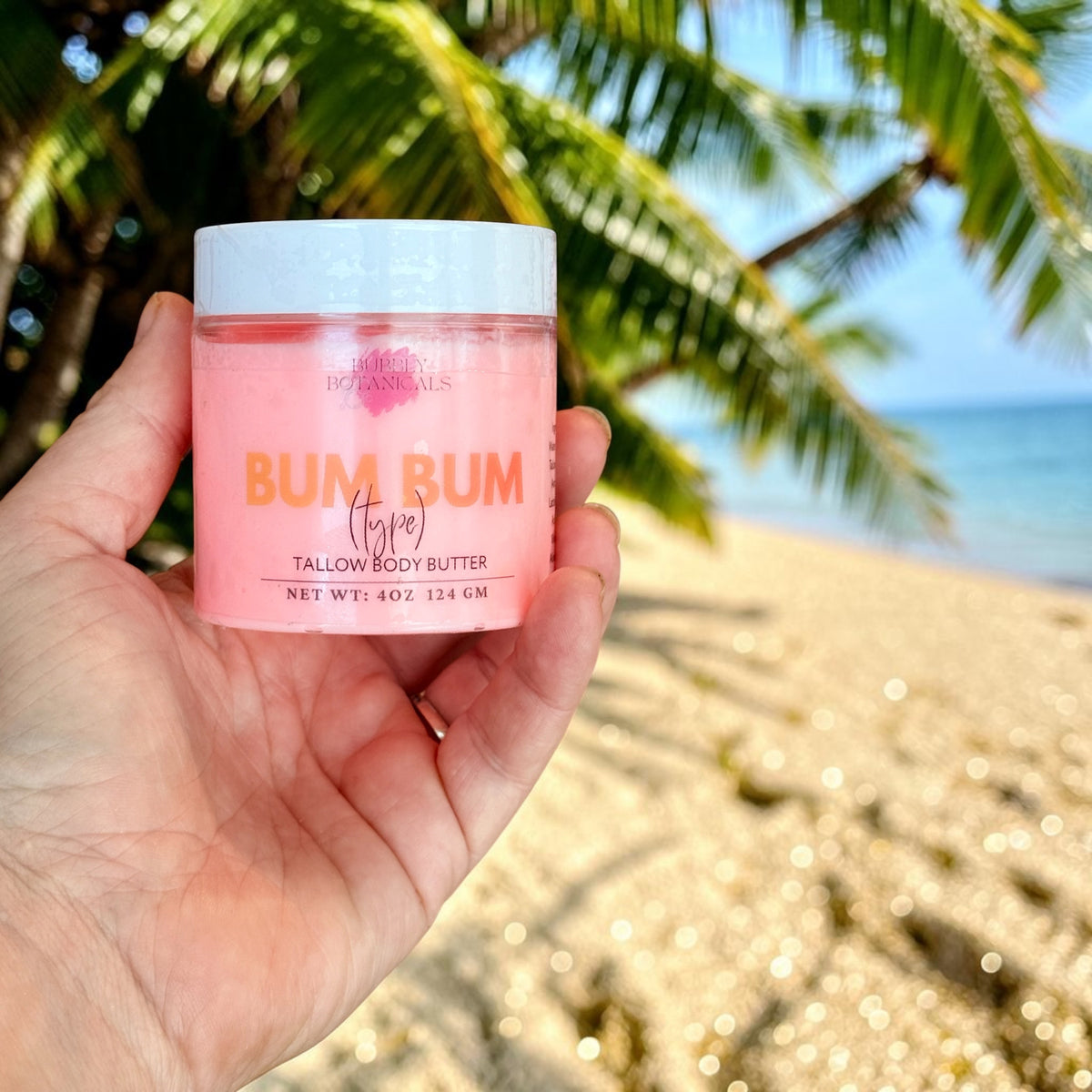 A hand holds a jar of Bum Bum Tallow Body Butter against a tropical beach background with palm trees and ocean water.