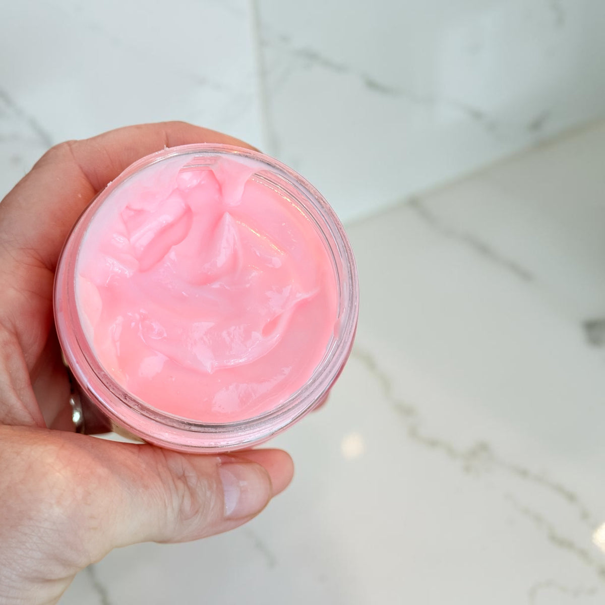 A hand holding an open clear jar filled with bright pink cream or balm, photographed over a light marble surface.