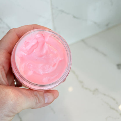 A hand holding an open clear jar filled with bright pink cream or balm, photographed over a light marble surface.