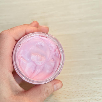 A hand holding a small clear jar filled with pale pink sweet Grace type tallow body butter, photographed from above against a elight wood surface.