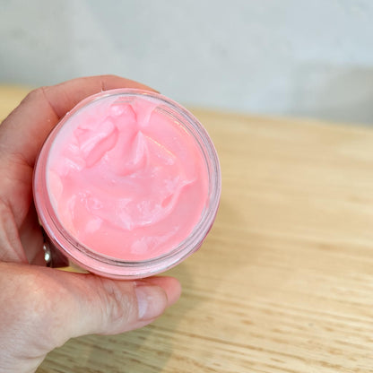 A hand holding an open clear jar filled with pink tallow body butter, photographed over a light wood tabletop with a soft blurred background.