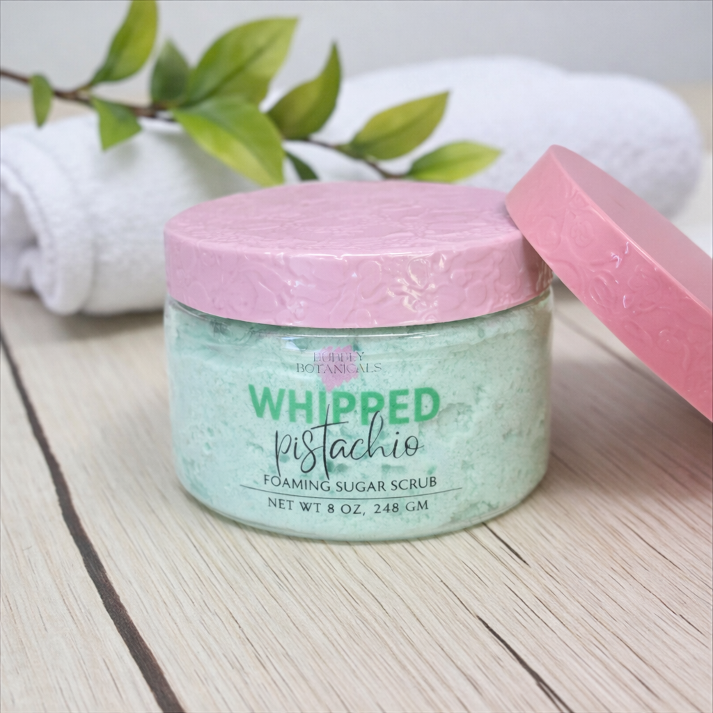 Jar of 'Whipped Pistachio' foaming sugar scrub with pink lid on a wooden surface.