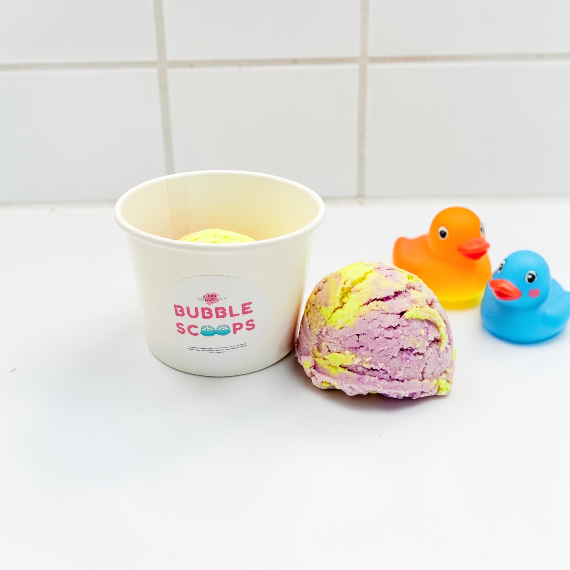 Bath bomb with a container labeled 'Bubble Scoops' and two rubber duck toys on a tiled bathroom floor.